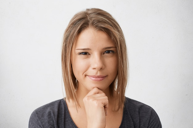 Nahaufnahmeporträt der niedlichen jungen Frau mit der trendigen Frisur und der reinen Haut, die lässige Kleidung trägt, die Hand auf Wange hält, die verdächtiges Aufstellen schaut.