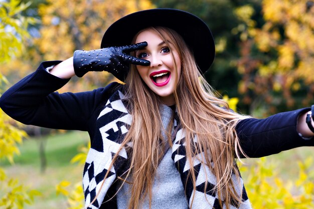 Nahaufnahmeporträt der prächtigen Dame im schwarzen Hut, der während des Herbst-Fotoshootings herumalbert. Lustige junge Dame in eleganten Handschuhen, die Zeit im Park am Septembertag verbringen.