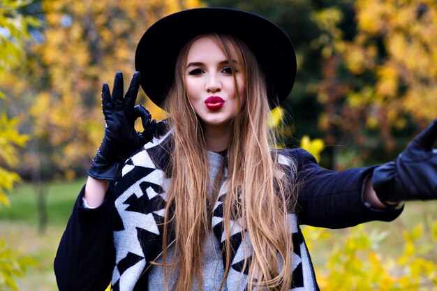 Nahaufnahmeporträt der prächtigen Dame im schwarzen Hut, der während des Herbst-Fotoshootings herumalbert. Lustige junge Dame in eleganten Handschuhen, die Zeit im Park am Septembertag verbringen.