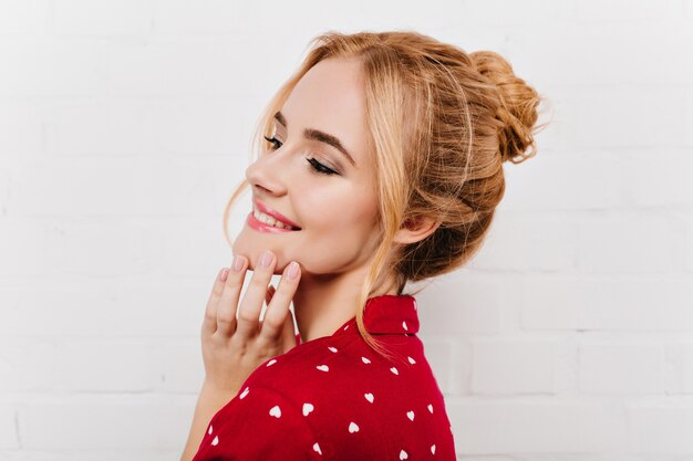 Nahaufnahmeporträt der reizenden blonden Frau mit niedlichem Make-up, das auf weißer Wand aufwirft. Schönes Mädchen mit stilvoller Frisur, die sanft ihr Gesicht mit einem Lächeln berührt.