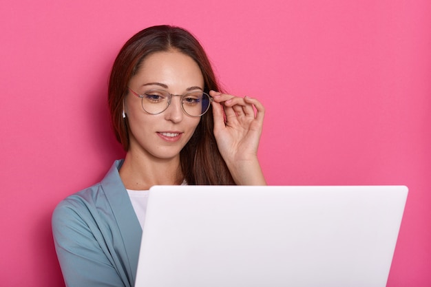 Nahaufnahmeporträt der reizenden Dame, die Rahmen ihrer Brille mit Hand berührt und Laptop-Bildschirm betrachtet