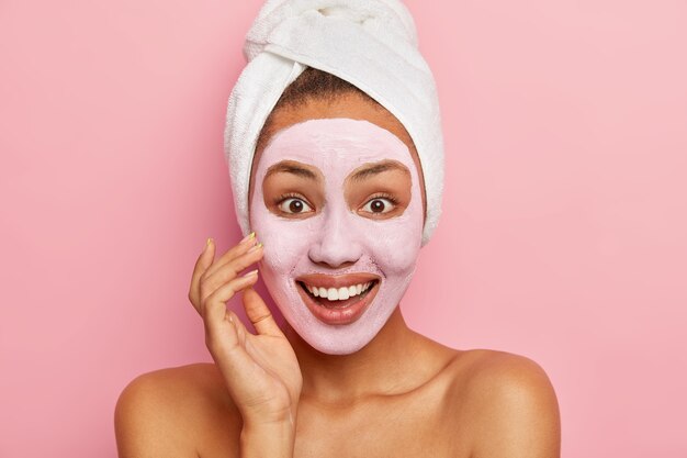 Nahaufnahmeporträt der reizenden glücklichen jungen Frau trägt Gesichtsmaske auf Teint auf, verwendet pflegenden Ton zur Verjüngung, trägt eingewickeltes weißes Handtuch auf Kopf