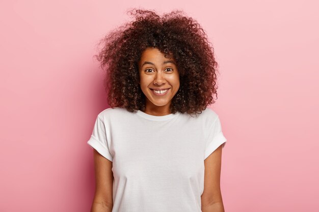 Nahaufnahmeporträt der reizenden jungen afroamerikanischen Frau lächelt positiv, genießt lustige Szene, amüsiert sich, kichert über lustigen Witz, hat dunkles lockiges voluminöses Haar, trägt lässiges Outfit