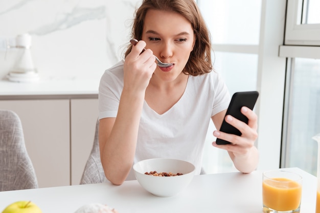 Nahaufnahmeporträt der schönen Frau, die auf Handy während des Essens Cornflakes am Küchentisch chattet