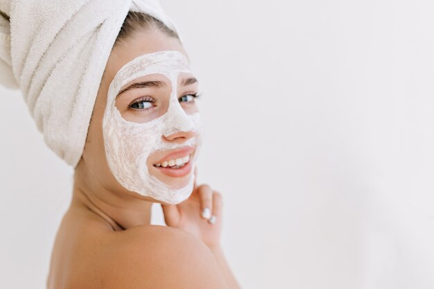 Nahaufnahmeporträt der schönen jungen Frau, die mit Handtüchern nach dem Bad lächelt, machen kosmetische Maske auf ihrem Gesicht.