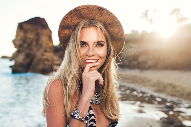 Nahaufnahmeporträt des attraktiven blonden Mädchens mit dem langen Haar, das auf felsigem Strand auf Sonnenunterganghintergrund aufwirft. Sie hält Finger auf Lippen und lächelt zur Kamera.