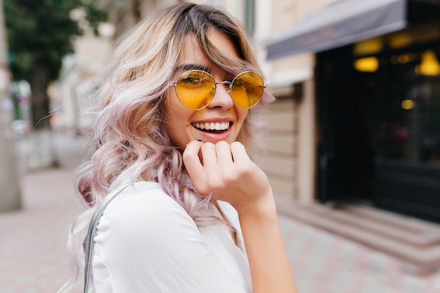 Nahaufnahmeporträt des attraktiven freudigen Mädchens mit erstaunlichem Lächeln trägt stilvolle gelbe Sonnenbrille und weißes Hemd