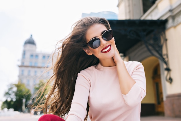 Nahaufnahmeporträt des attraktiven Mädchens in der Sonnenbrille mit weinigen Lippen in der Stadt. Ihr langes Haar fliegt im Wind, sie lächelt mit schneeweißem Lächeln.