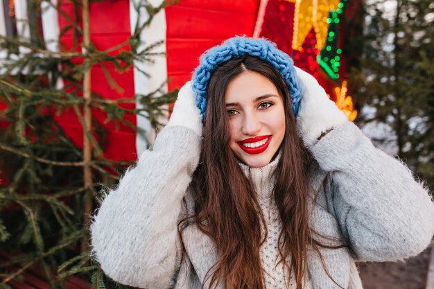 Nahaufnahmeporträt des begeisterten Mädchens im blauen Hut, das mit glücklichem Gesichtsausdruck vor Weihnachtsbäumen aufwirft. Foto im Freien der glamourösen Frau mit dunklem Haar, das nahe Neujahrsdekoration steht.