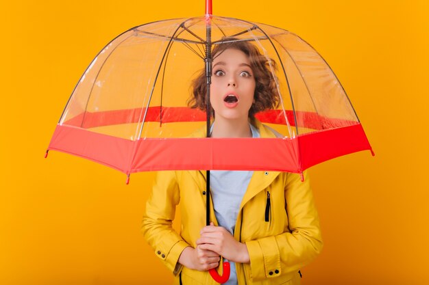 Nahaufnahmeporträt des begeisterten Mädchens mit der lockigen Frisur, die unter Sonnenschirm steht. Innenfoto des verärgerten weiblichen Modells im Regenmantel, der Regenschirm hält.