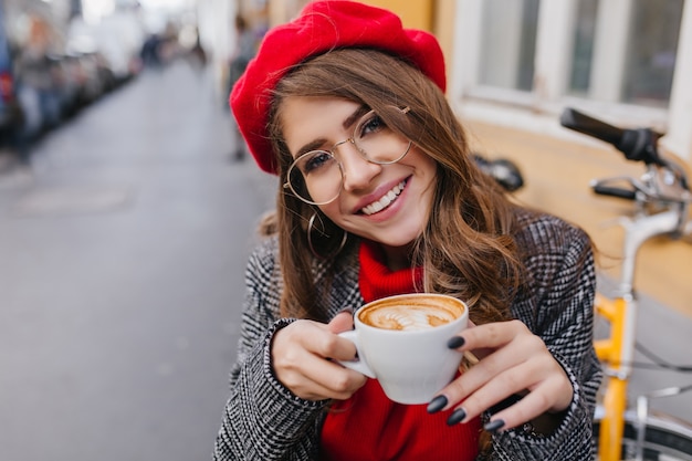 Nahaufnahmeporträt des blassen brünetten Mädchens mit dem charmanten Lächeln, das sanft Tasse Kaffee hält