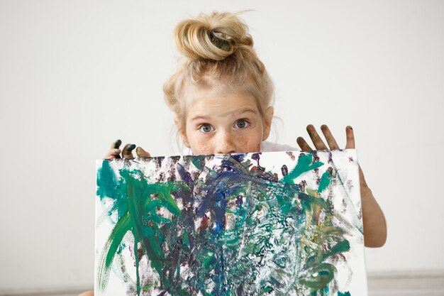 Nahaufnahmeporträt des blonden europäischen kleinen Mädchens mit Haarknoten und großen blauen Augen, die ihr Bild demonstrieren.