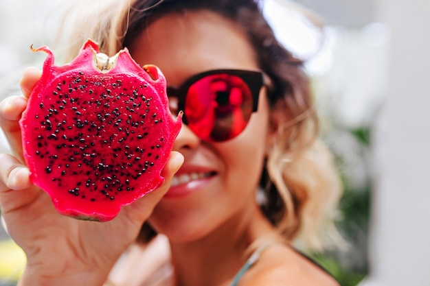 Nahaufnahmeporträt des blonden Mädchens in den hellen Gläsern, die mit pitaya aufwerfen. Winsome kaukasische Dame, die exotische Frucht hält.