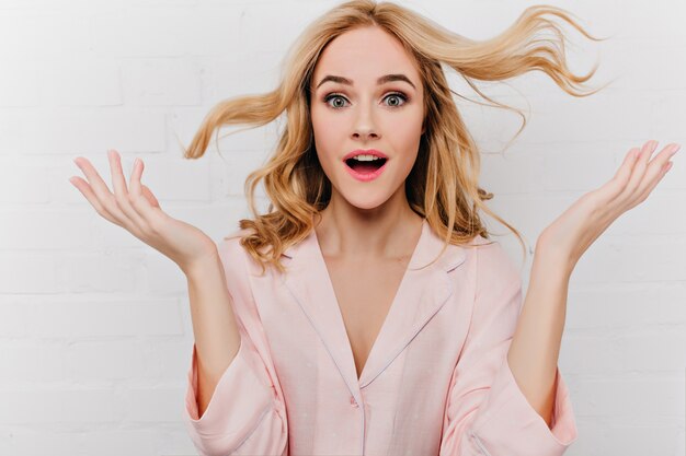 Nahaufnahmeporträt des blonden Mädchens in der rosa Kleidung, die Erstaunen auf weißer Wand ausdrückt. Überraschte blonde Dame im Nachtanzug posiert emotional