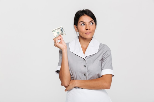 Nahaufnahmeporträt des denkens der hübschen brünetten frau in der grauen uniform, die dollarbanknote hält und beiseite schaut