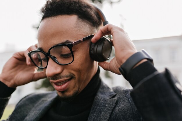 Nahaufnahmeporträt des entzückenden schwarzen Mannes mit stilvollem Haarschnitt, der Musik mit geschlossenen Augen hört. Foto des müden afrikanischen Kerls in den Gläsern, die Lied in Kopfhörern genießen.