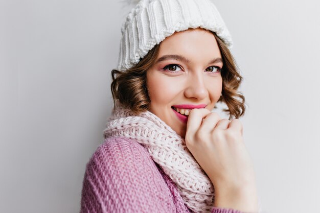 Nahaufnahmeporträt des erstaunlichen europäischen Mädchens im lila Wollpullover, der mit Lächeln aufwirft. Foto der eleganten dunkelhaarigen Dame in der Mütze und im Schal.