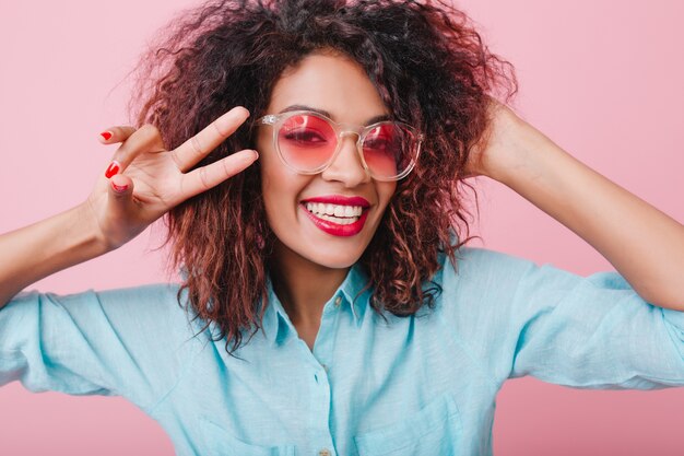 Nahaufnahmeporträt des fröhlichen afrikanischen Mädchens, das wahres Glück ausdrückt. hübsche Mulattendame in der rosa Brille, die Friedenszeichen zeigt.