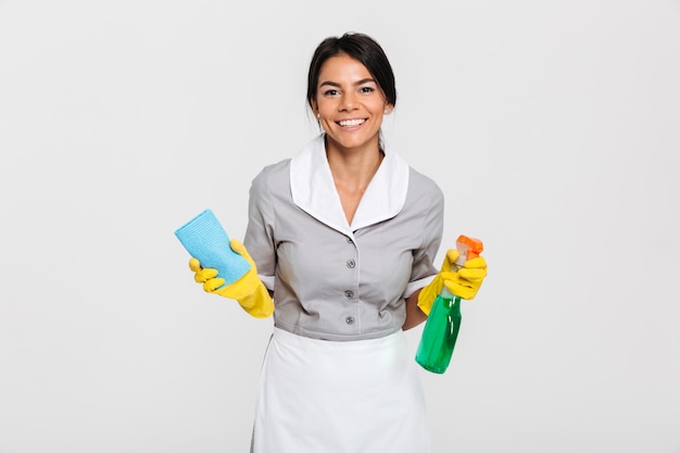 Nahaufnahmeporträt des fröhlichen Dienstmädchens in der Uniform, die Lappen und Reinigungsspray hält