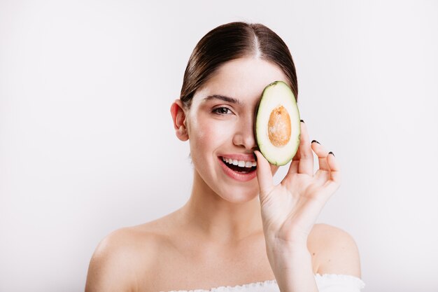 Nahaufnahmeporträt des gesunden Mädchens mit der sauberen strahlenden Haut, die mit Avocado auf weißer Wand aufwirft.