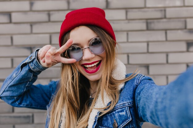 Nahaufnahmeporträt des glückseligen weißen Mädchens, das Spaß im Freien hat. Nettes blondes weibliches Modell, das tanzt und Selfie auf Backsteinmauer macht.