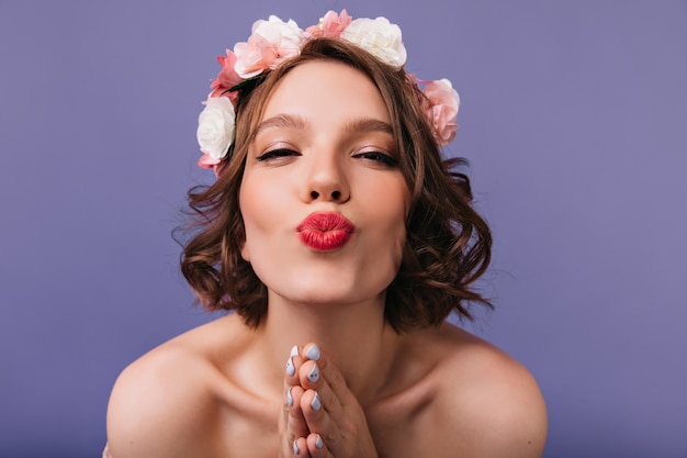 Nahaufnahmeporträt des glückseligen weißen Mädchens mit rosa Rosen im Haar. Atemberaubende Frau trägt einen Blumenstrauß, der mit küssendem Gesichtsausdruck aufwirft.