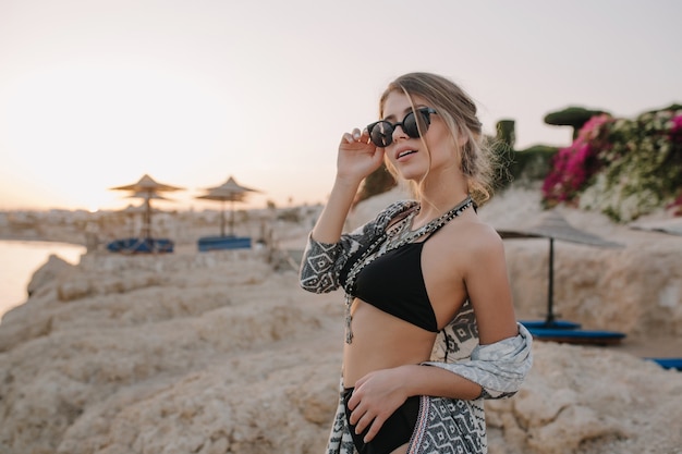 Nahaufnahmeporträt des herrlichen jungen Mädchens mit dem schlanken Körper, der stilvollen schwarzen Badeanzug, Halskette, Strickjacke, Umhang mit Verzierungen trägt. Strand, Felsen, Sonnenuntergang.