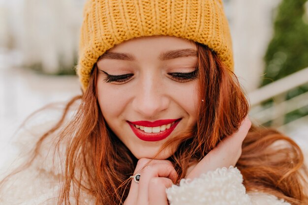 Nahaufnahmeporträt des herrlichen Mädchens mit den roten Lippen. Schüchterne Ingwerfrau im Hut, der im Winter aufwirft.