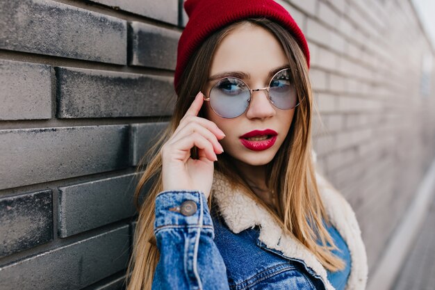 Nahaufnahmeporträt des herrlichen weißen Mädchens im Frühlingsoutfit, das auf der Straße aufwirft. Interessierte kaukasische Frau trägt Jeansjacke und roten Hut, der auf städtischer Straße steht.