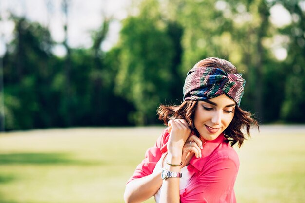 Nahaufnahmeporträt des hübschen Brunettemädchens im Bandana, das im Sommerpark geht. Sie trägt weiße Kleidung, langes rosa Hemd. Sie lächelt nach unten.