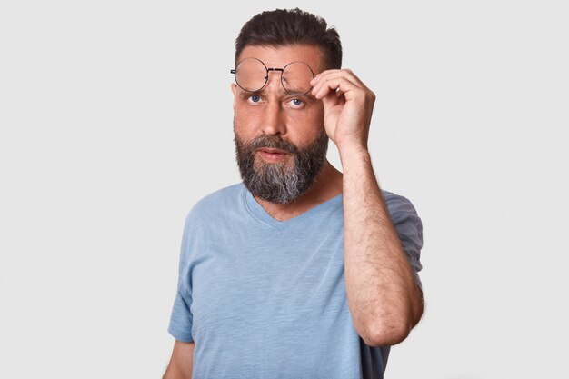 Nahaufnahmeporträt des hübschen jungen bärtigen Mannes mit Brille, der Freizeitkleidung trägt, auf Weiß steht, seine Brille fragend anhebt, Zweifel und.