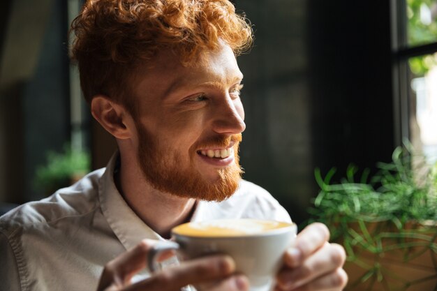 Nahaufnahmeporträt des hübschen lächelnden bärtigen rothaarigen Mannes, der Kaffeetasse hält und beiseite schaut