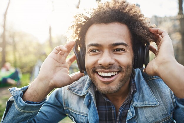 Nahaufnahmeporträt des hübschen unrasierten dunkelhäutigen Kerls, der Hände auf Kopfhörern hält, während Musik hört und aufgeregt ist, beiseite schaut, im Park sitzt.