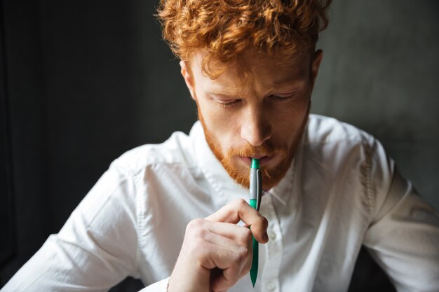 Nahaufnahmeporträt des jungen konzentrierten gelockten Lesekopfmanns mit Stift in seinem Mund