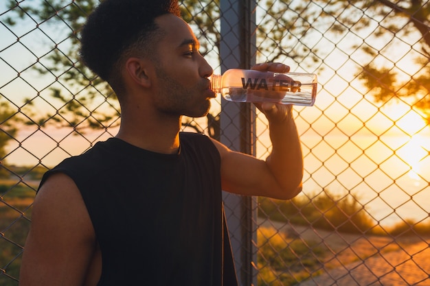 Nahaufnahmeporträt des jungen Mannes, der morgens Sport treibt, Wasser auf dem Basketballplatz bei Sonnenaufgang trinkt