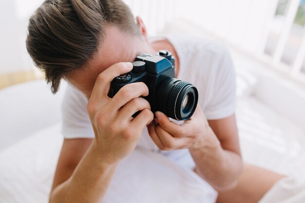 Nahaufnahmeporträt des jungen Mannes mit der trendigen Frisur, die professionelle Kamera in den Händen hält. Kerl mit dem dunklen kurzen Haar im weißen Hemd, das neue Fotos für Portfolio am sonnigen Morgen macht