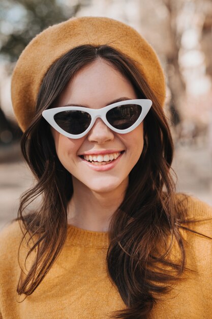 Nahaufnahmeporträt des lächelnden Mädchens in der gelben Baskenmütze und in der Sonnenbrille. Brünette junge Frau im orangefarbenen Pullover lachend beim Gehen