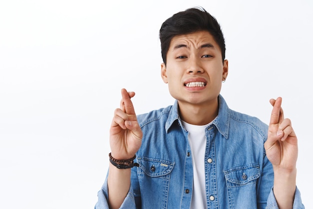 Nahaufnahmeporträt des nervösen jungen asiatischen Mannes, der besorgt über Ergebnisse ist, kreuzt die Finger, viel Glück, presst die Zähne zusammen und schaut hoffnungsvoll in die Kamera, die um Traum bittet, wird wahr und plädiert auf weißem Hintergrund