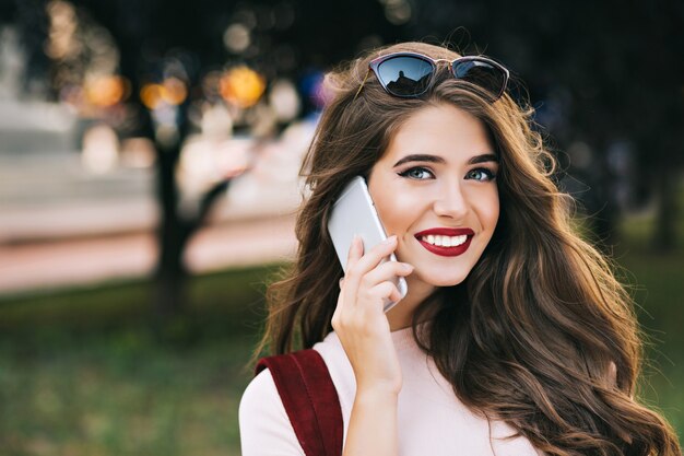 Nahaufnahmeporträt des niedlichen Mädchens mit effektivem Makeaup und langen Haaren, die am Telefon im Park sprechen. Sie hat weinige Lippen und lächelt.