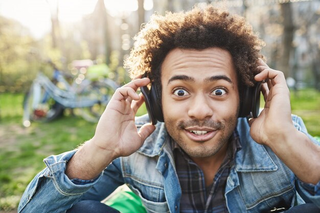 Nahaufnahmeporträt des niedlichen modischen Afroamerikaners, der mit geknallten Augen schaut und Augenbrauen an der Kamera anhebt, während er im Park sitzt und Musik über Kopfhörer hört und Aufregung ausdrückt