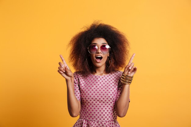 Nahaufnahmeporträt des Retro stilvollen verblüfften afrikanischen Frauen in der Sonnenbrille, die mit zwei Fingern nach oben zeigt und nach oben schaut