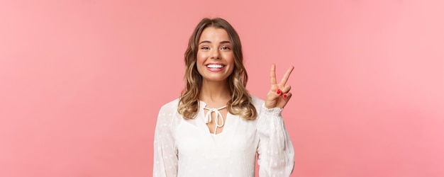 Nahaufnahmeporträt des romantischen reizenden lächelnden Mädchens mit dem blonden kurzen Haar, das weißes Kleid trägt, zeigen Friedenszeichen, das Frühlingsgrinsen genießt und Kamera über rosa Hintergrund schaut