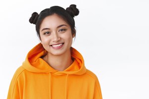Nahaufnahmeporträt des schönen asiatischen mädchens mit den netten zwei hairbuns, die den orange stilvollen hoodie tragen, der glücklich lächelt, drücken positive gefühle konzept des schönheitsmake-ups und des weißen hintergrundes der mode aus