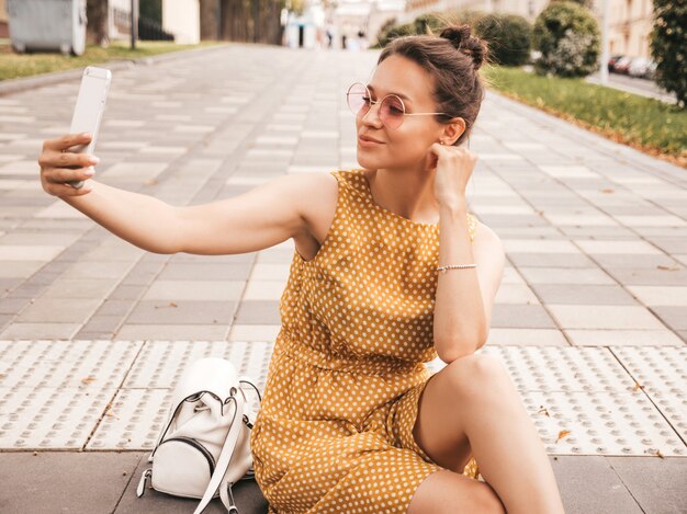 Nahaufnahmeporträt des schönen lächelnden Brunettemädchens im Sommerhippie-Gelbkleid. Vorbildliches nehmendes selfie auf Smartphone Frau, die Fotos am warmen sonnigen Tag in der Straße in der Sonnenbrille macht