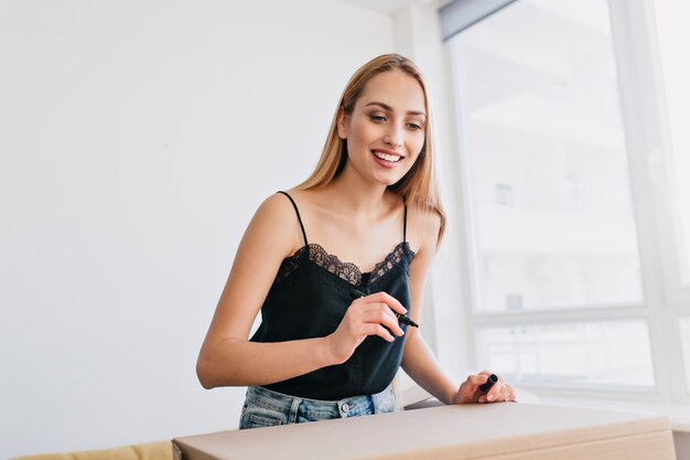 Nahaufnahmeporträt des schönen Mädchens, das Pappkarton, Verpackung, Umzug in neue Wohnung, Haus beschriftet. Lächelnde Dame im weißen Raum mit Fenster, schwarzes Oberteil und Jeans tragend.