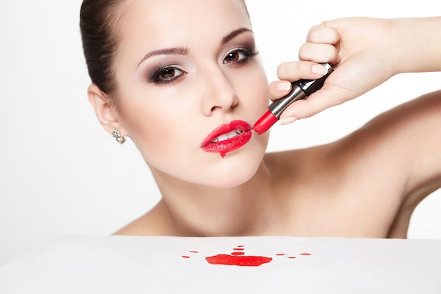 Nahaufnahmeporträt des sexy kaukasischen jungen Frauenmodells mit glamourösen roten Lippen, hellem Make-up, Augenpfeil-Make-up, reinem Teint mit rotem Lippenstift. Perfekt saubere Haut. Blut auf dem Tisch