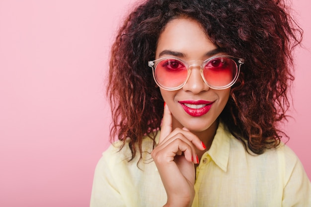 Nahaufnahmeporträt des sinnlich interessierten Mädchens in der rosa Brille, die Wange mit Hand berührt. Liebenswerte Dame mit brauner Haut und strahlendem Make-up lächelnd.