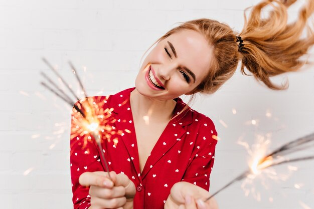 Nahaufnahmeporträt des sorglosen Mädchens lokalisiert auf weißer Wand mit bengalischen Lichtern. emotionale Dame im roten Nachtanzug posiert mit Wunderkerzen vor Neujahr.