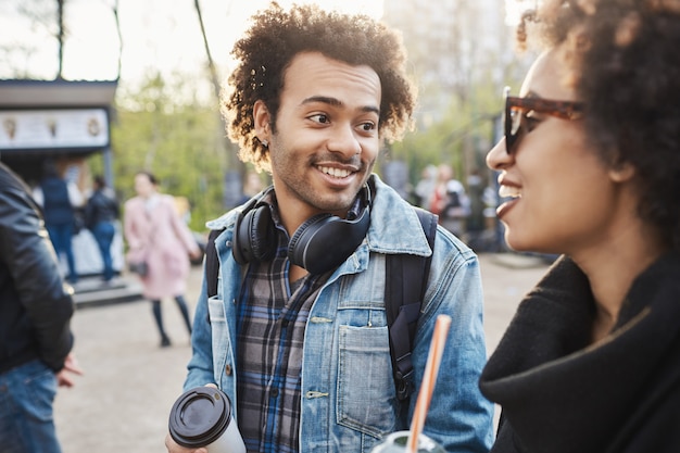 Nahaufnahmeporträt des stilvollen Afroamerikaners mit niedlichem Lächeln und Afro-Frisur, die mit Geschwister sprechen, während sie Spaß im Park haben, Kaffee trinken und herum scherzen.