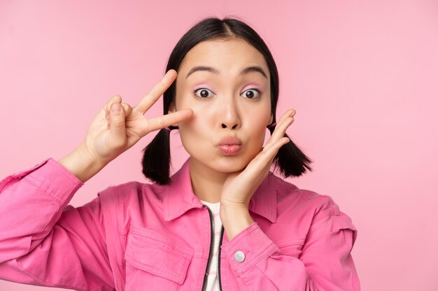 Nahaufnahmeporträt des stilvollen asiatischen Mädchens zeigt Friedenszeichen und küsst sich vor rosa Hintergrund